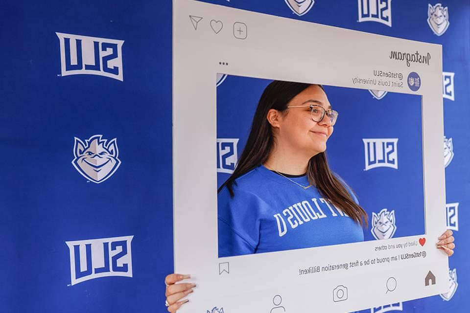 A student wearing a 博彩网址大全 sweatshirt stands in front of a 博彩网址大全 banner while holding up a picture frame that says First Generation at 博彩网址大全 and Proud to be a first-generation Billiken